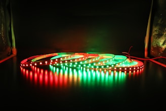 A series of LED light strips are coiled on a dark surface, illuminated in alternating red and green colors. The glowing lights create reflections on the glossy surface below, providing a striking contrast against the dark background.