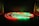 A series of LED light strips are coiled on a dark surface, illuminated in alternating red and green colors. The glowing lights create reflections on the glossy surface below, providing a striking contrast against the dark background.