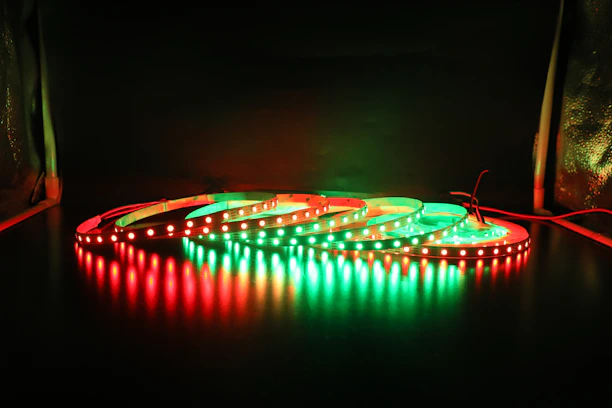 A series of LED light strips are coiled on a dark surface, illuminated in alternating red and green colors. The glowing lights create reflections on the glossy surface below, providing a striking contrast against the dark background.