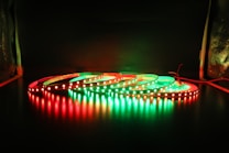A series of LED light strips are coiled on a dark surface, illuminated in alternating red and green colors. The glowing lights create reflections on the glossy surface below, providing a striking contrast against the dark background.