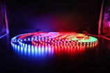 A coiled strip of LED lights lies on a reflective surface, emitting bright colors of blue, red, and green. The colorful lights are vividly illuminated against a dark background, creating a striking contrast.