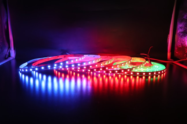 A coiled strip of LED lights lies on a reflective surface, emitting bright colors of blue, red, and green. The colorful lights are vividly illuminated against a dark background, creating a striking contrast.