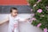 A young child wearing a light-colored onesie reaches out to touch a branch with vibrant pink flowers. The scene is set against a background of a wall with a red and gray color scheme. A hand gently supports the child, ensuring they remain steady.