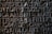 A detailed view of a textured, metallic surface with raised letters forming words and phrases. The text appears to be sculpted or engraved into the metal with an intricate design.