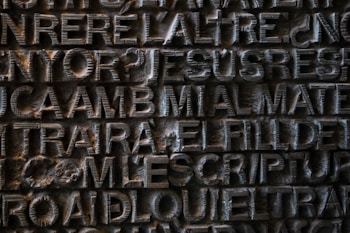 A detailed view of a textured, metallic surface with raised letters forming words and phrases. The text appears to be sculpted or engraved into the metal with an intricate design.