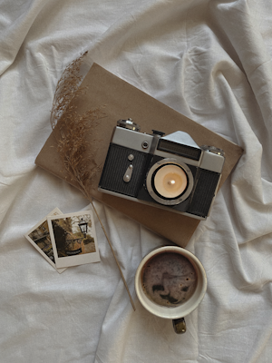 A vintage camera with a lit candle inside the lens is placed on a brown hardcover book. Two Polaroid photos lie beside the camera, and nearby is a cup of coffee with foam. There is also a dried sprig of grass. The items are arranged on a soft, wrinkled white or light grey fabric.