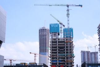 A construction site with multiple high-rise buildings in various stages of completion. Large cranes are positioned around the structures, indicating ongoing development. The buildings are surrounded by a bright blue sky with scattered clouds, suggesting a clear day.