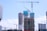 A construction site with multiple high-rise buildings in various stages of completion. Large cranes are positioned around the structures, indicating ongoing development. The buildings are surrounded by a bright blue sky with scattered clouds, suggesting a clear day.