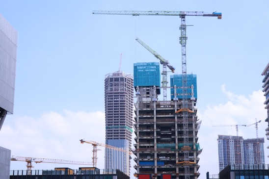 A construction site with multiple high-rise buildings in various stages of completion. Large cranes are positioned around the structures, indicating ongoing development. The buildings are surrounded by a bright blue sky with scattered clouds, suggesting a clear day.