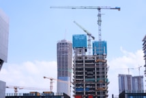 A construction site with multiple high-rise buildings in various stages of completion. Large cranes are positioned around the structures, indicating ongoing development. The buildings are surrounded by a bright blue sky with scattered clouds, suggesting a clear day.