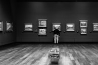 A person is observing a collection of framed paintings in an art gallery. The gallery has a wooden floor and muted wall colors, creating a serene and reflective atmosphere. The paintings are evenly spaced on the walls, and the viewer stands at a comfortable distance while appreciating the art.