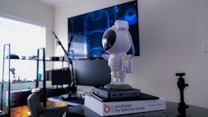 An office or study space features a small astronaut figurine with a reflective helmet atop a closed laptop, which is placed on a book titled 'JavaScript: The Definitive Guide.' In the background, computer monitors display a schematic resembling Iron Man's helmet, and shelves filled with books and various objects are visible. A soft light enters from a window partially covered by blinds, creating a tranquil atmosphere.