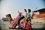 A man and a woman are sitting on a decorated camel on a sandy beach. The woman is taking a selfie with a phone, while the man smiles. The camel is adorned with colorful tassels in red, pink, and blue. In the background, there are buildings and a clear blue sky.