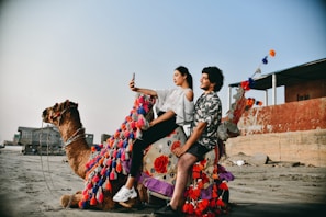 A man and a woman are sitting on a decorated camel on a sandy beach. The woman is taking a selfie with a phone, while the man smiles. The camel is adorned with colorful tassels in red, pink, and blue. In the background, there are buildings and a clear blue sky.