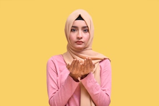 A person wearing a light brown hijab and pink sweater holds their hands up, palms facing up, in a prayer-like gesture. Henna designs are visible on the palms. The background is a solid yellow color.