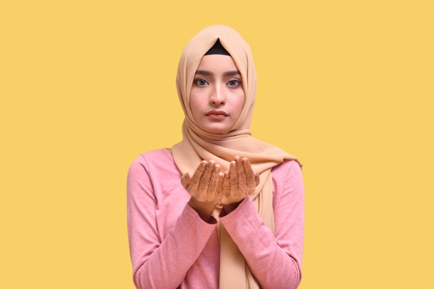 A person wearing a light brown hijab and pink sweater holds their hands up, palms facing up, in a prayer-like gesture. Henna designs are visible on the palms. The background is a solid yellow color.