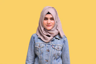 A graceful woman in a flowing beige abaya and soft cream hijab, standing against a minimalist white backdrop with gentle gold accents.