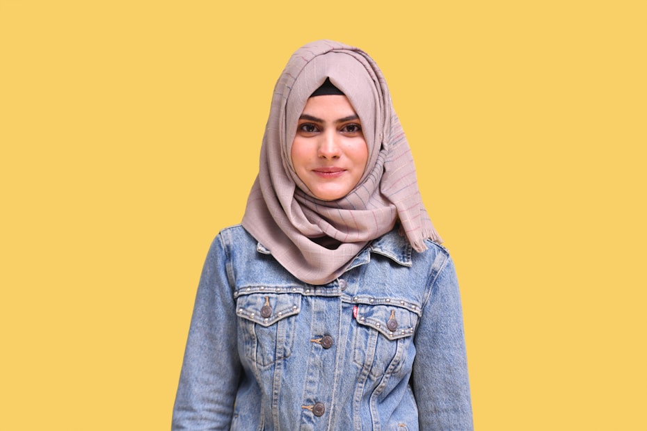 A person wearing a beige hijab and a denim jacket is standing against a plain yellow background.