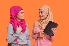 Two women are standing and facing each other, smiling. One woman is wearing a bright pink hijab and the other is wearing a beige hijab. The woman in the beige hijab is holding a black notebook. Both appear to be engaged in a friendly conversation.