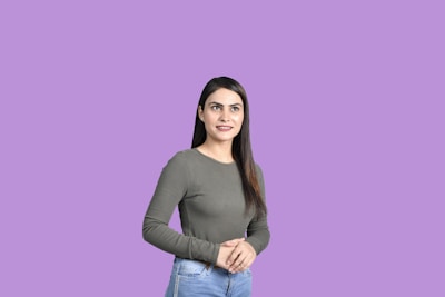 A woman with long dark hair wearing a gray long-sleeve top and jeans stands against a solid lavender background. She has a calm expression and her hands are gently clasped in front of her.