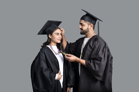 Two people wearing graduation caps and gowns. One person is adjusting the tassel on the other's cap. Both are smiling slightly, wearing medals around their necks.