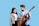 A young man and woman are standing together with the man holding an acoustic guitar. Both are smiling, and the woman is looking at the guitar. The man is casually dressed in a denim shirt, while the woman wears a grey jacket over a pink t-shirt.