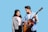A young man and woman are standing together with the man holding an acoustic guitar. Both are smiling, and the woman is looking at the guitar. The man is casually dressed in a denim shirt, while the woman wears a grey jacket over a pink t-shirt.