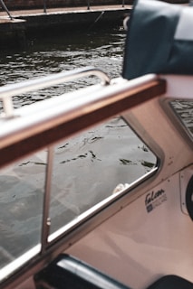 A portion of a boat interior with visible upholstery and a wooden armrest, partially overlooking a body of water with gentle ripples. The water is bordered by a concrete edge and metal railing.