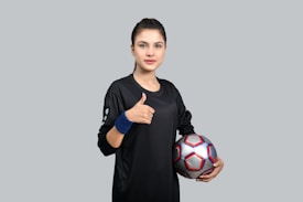 A person in a black sports jersey holds a soccer ball under one arm while giving a thumbs-up with the other hand. The background is a plain gray color, and the person is wearing a blue wristband.