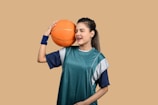 A young woman wearing a sports jersey and wristband holds a basketball against her head with one hand, eyes closed and displaying a relaxed expression. The background is a solid tan color.