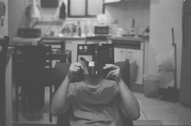 A person is seated in a cozy kitchen area, holding a book that covers their face. The kitchen has a soft, cluttered appearance with everyday items like a dish rack, bottles, and a mop in view.