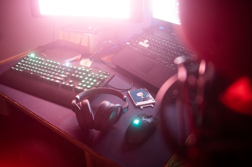 A dimly lit desk with a mechanical keyboard featuring illuminated keys, a pair of headphones, a laptop, and a mouse with glowing accents. A hard drive and an open notebook are also present. The overall setup gives a gaming or tech vibe.