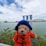 A plush teddy bear wearing a blue hat and a red coat stands in front of a body of water. In the background, the iconic Marina Bay Sands hotel and the Singapore Flyer are visible, along with the ArtScience Museum. The sky is partly cloudy, creating a picturesque scene.