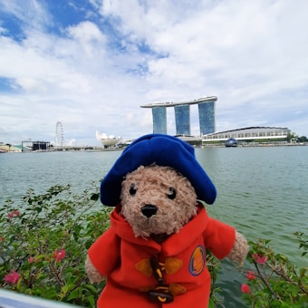 A plush teddy bear wearing a blue hat and a red coat stands in front of a body of water. In the background, the iconic Marina Bay Sands hotel and the Singapore Flyer are visible, along with the ArtScience Museum. The sky is partly cloudy, creating a picturesque scene.