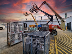 A construction site at dusk with a vibrant, colorful sky. Two aerial work platforms are visible, with one in the foreground and one elevated at the back. The area is marked with yellow safety lines and there are metal cages containing gas cylinders. A truck is parked nearby, and there are industrial buildings in the background.