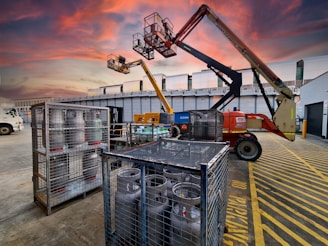 A construction site at dusk with a vibrant, colorful sky. Two aerial work platforms are visible, with one in the foreground and one elevated at the back. The area is marked with yellow safety lines and there are metal cages containing gas cylinders. A truck is parked nearby, and there are industrial buildings in the background.