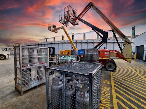 A construction site at dusk with a vibrant, colorful sky. Two aerial work platforms are visible, with one in the foreground and one elevated at the back. The area is marked with yellow safety lines and there are metal cages containing gas cylinders. A truck is parked nearby, and there are industrial buildings in the background.