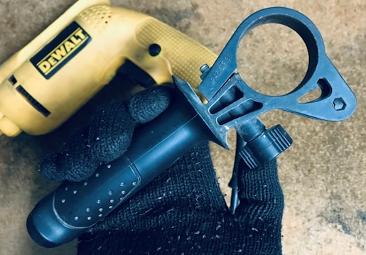 A close-up view of a black gloved hand holding a black, circular power tool attachment against a textured surface. In the background, there is a yellow power drill with a visible brand name.