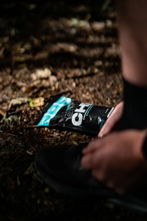 A person is tying the laces of a black shoe on a dirt or forest floor, with a black and teal packaging of an energy gel pack lying nearby. The scene is illuminated by dappled sunlight filtering through the trees.