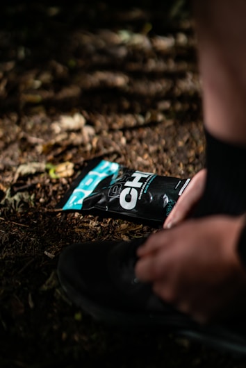 A person is tying the laces of a black shoe on a dirt or forest floor, with a black and teal packaging of an energy gel pack lying nearby. The scene is illuminated by dappled sunlight filtering through the trees.