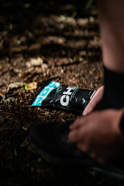 A person is tying the laces of a black shoe on a dirt or forest floor, with a black and teal packaging of an energy gel pack lying nearby. The scene is illuminated by dappled sunlight filtering through the trees.