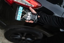 A hand holding a packaged product labeled 'CHU' is positioned near the rear light of a black car with a shiny surface. The product packaging features a combination of black, white, and turquoise colors. An arm dressed in a black long-sleeved garment extends towards the vehicle.