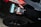 A hand holding a packaged product labeled 'CHU' is positioned near the rear light of a black car with a shiny surface. The product packaging features a combination of black, white, and turquoise colors. An arm dressed in a black long-sleeved garment extends towards the vehicle.