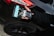 A hand holding a packaged product labeled 'CHU' is positioned near the rear light of a black car with a shiny surface. The product packaging features a combination of black, white, and turquoise colors. An arm dressed in a black long-sleeved garment extends towards the vehicle.