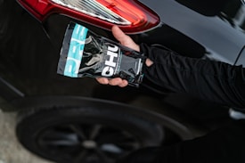 A hand holding a packaged product labeled 'CHU' is positioned near the rear light of a black car with a shiny surface. The product packaging features a combination of black, white, and turquoise colors. An arm dressed in a black long-sleeved garment extends towards the vehicle.