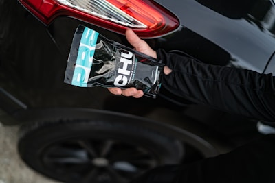 A hand holding a packaged product labeled 'CHU' is positioned near the rear light of a black car with a shiny surface. The product packaging features a combination of black, white, and turquoise colors. An arm dressed in a black long-sleeved garment extends towards the vehicle.