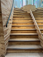 A wooden staircase with a black handrail ascends through a well-lit space. The steps are made of light-colored wood and are bordered by a tiled surface. Walls on either side are constructed from stacked wooden panels. Colorful stained glass windows are visible in the background, adding a vibrant touch to the setting.