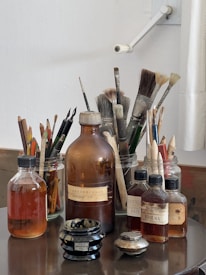 A collection of art supplies is neatly arranged on a table. Glass jars hold a variety of paintbrushes with different bristle sizes and shapes. There are several bottles containing liquids, possibly solvents or varnishes, and a small black jar labeled as cream. The background is a plain white wall with a mounted arm fixture.