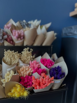 A collection of small bouquets arranged in brown paper wraps. The flowers are in a variety of vibrant colors, including yellow, pink, purple, red, and orange. They are displayed on black shelves against a deep blue background, adding to the contrast and vibrancy of the flowers.