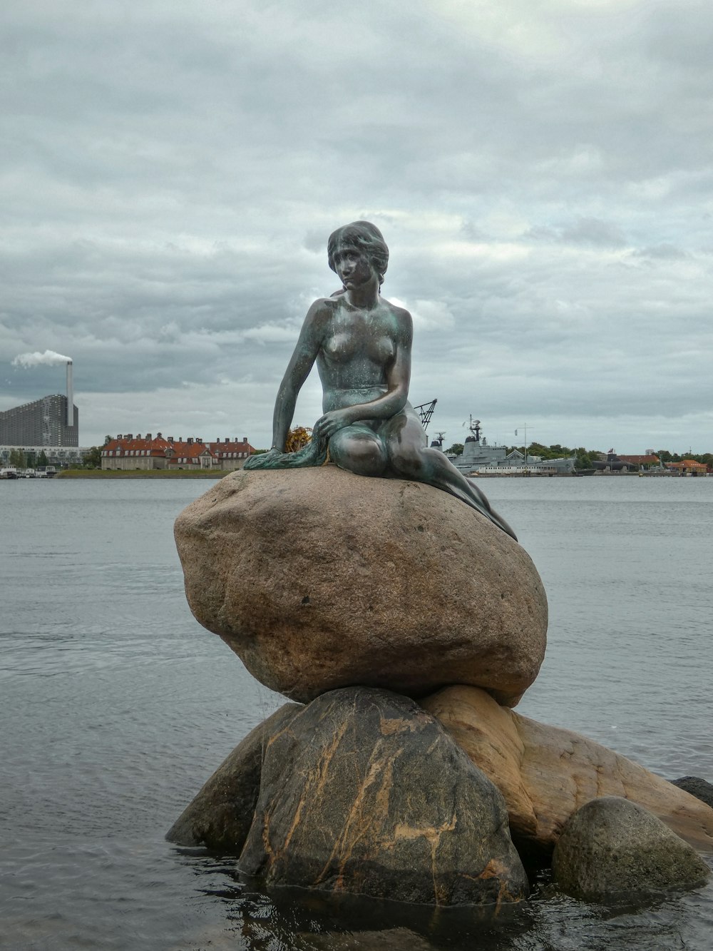 a statue of a man sitting on a rock in the water with The Little Mermaid in the background